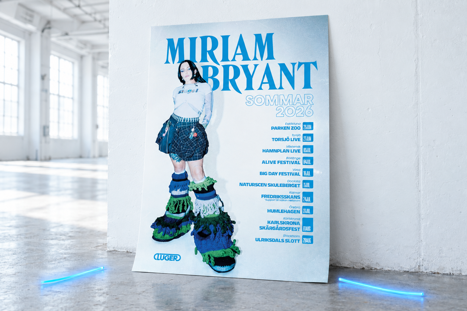 Miriam Bryant’s Sommar 2026 tour poster leaning against a white warehouse wall, with two curved blue glowsticks on the floor.